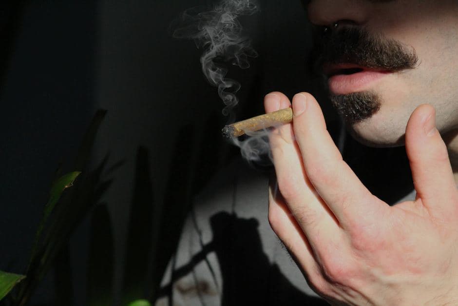 Een man met een snor houdt een smeulende joint met filter bij zijn mond terwijl er rook opstijgt.