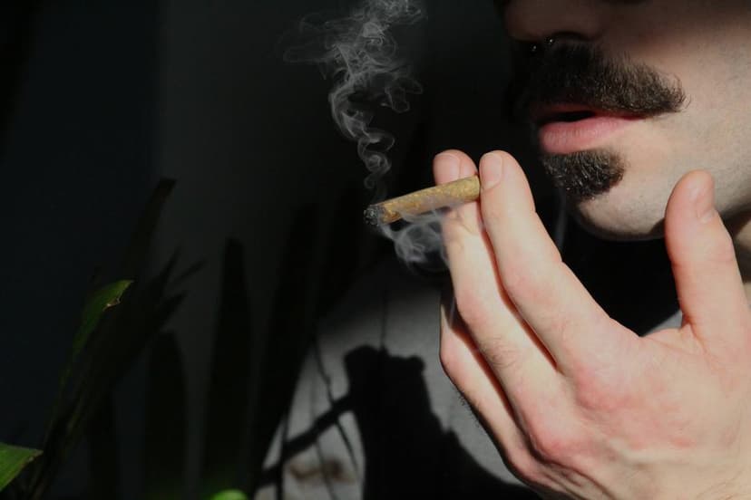 Een man met een snor houdt een smeulende joint met filter bij zijn mond terwijl er rook opstijgt.