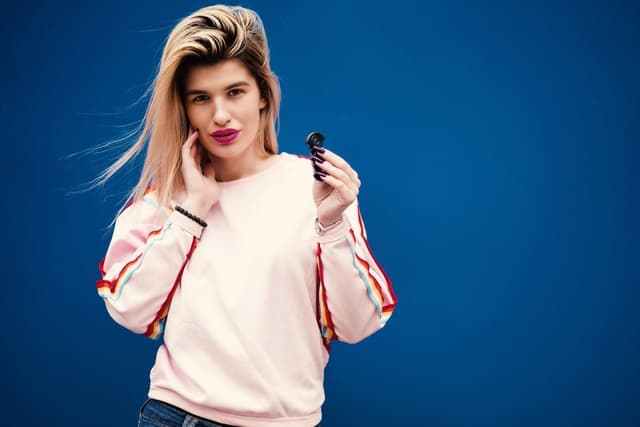 Een stijlvolle vrouw in een roze trui houdt een klein zwart bongonderdeel vast voor een blauwe achtergrond.