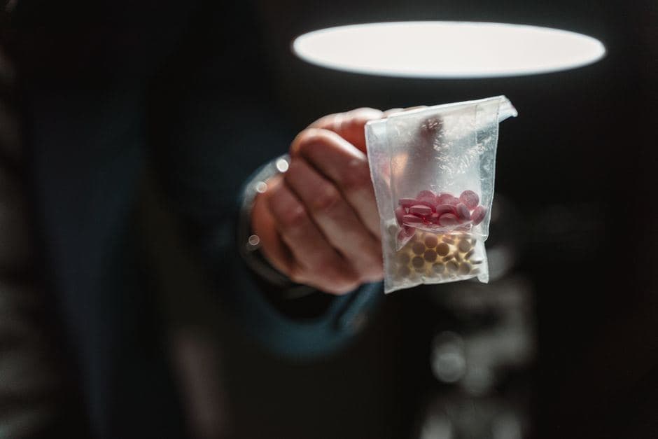 Een hand houdt in het donker een transparant plastic zakje met pillen omhoog onder een felle lamp.