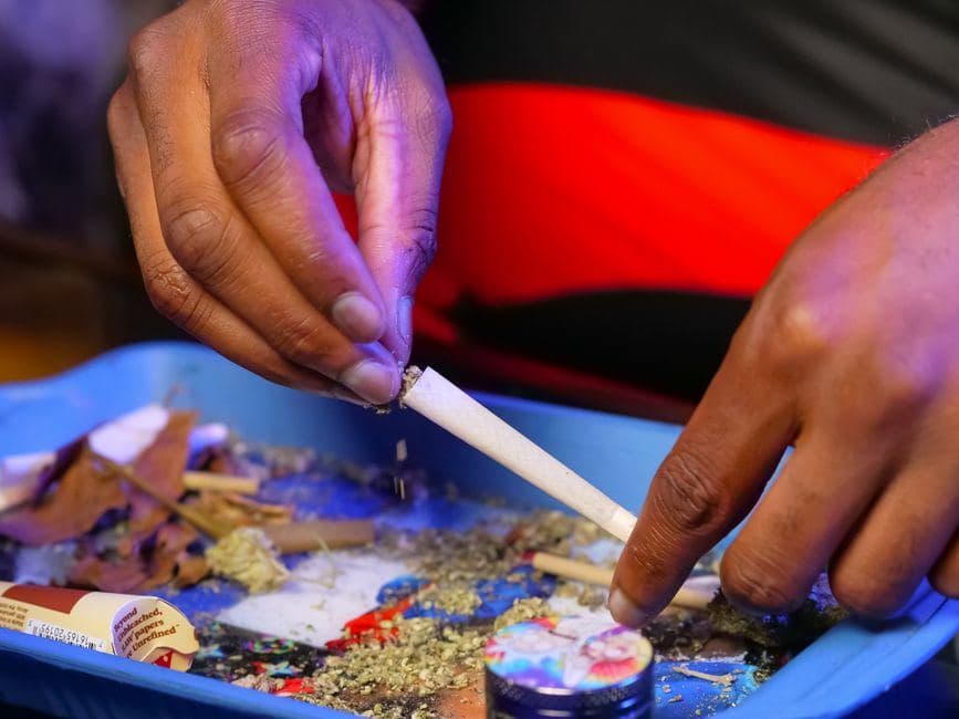 Handen die stap voor stap een joint draaien en vullen met cannabis boven een blauwe tray met een grinder.