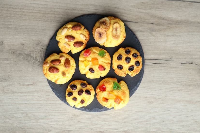 Zeven ronde mini-spacecakes versierd met amandelen, rozijnen, banaan en gekonfijt fruit op een donkere serveerplank.