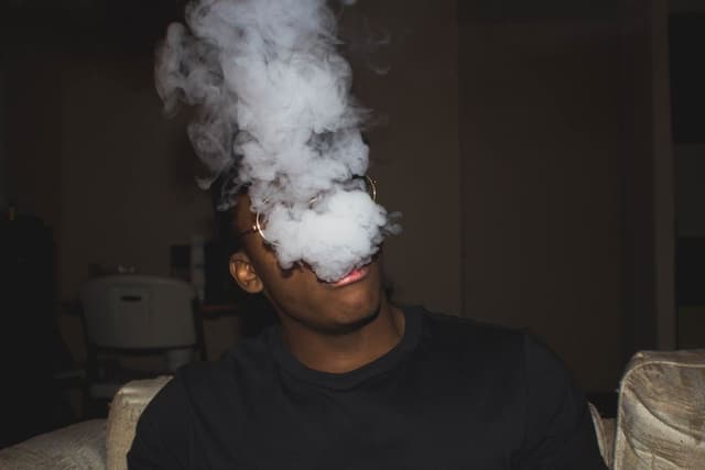 Man met bril blaast een dikke wolk damp uit die zijn gezicht bedekt, zittend op een bank