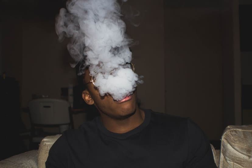 Man met bril blaast een dikke wolk damp uit die zijn gezicht bedekt, zittend op een bank