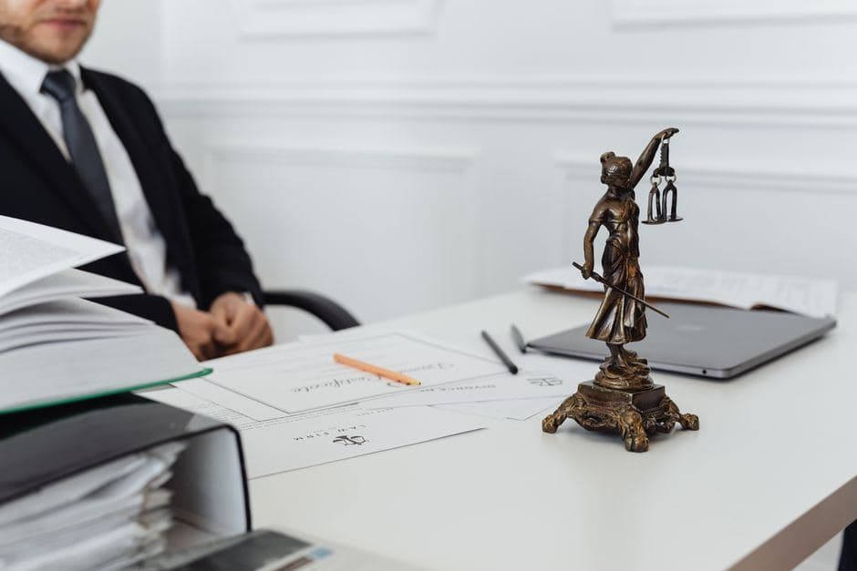 Een advocaat in pak zit achter een bureau met juridische documenten, boeken en een beeldje van Vrouwe Justitia.