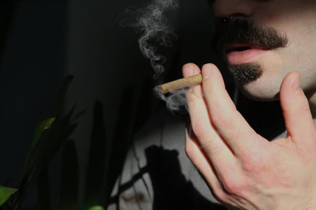 Een man met een snor houdt een smeulende joint met filter bij zijn mond terwijl er rook opstijgt.