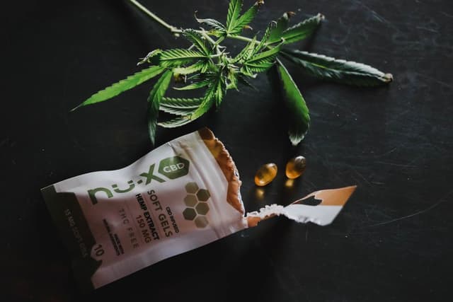 Een geopend zakje CBD-softgels met twee goudgele capsules en een hennepblad op een zwarte ondergrond.