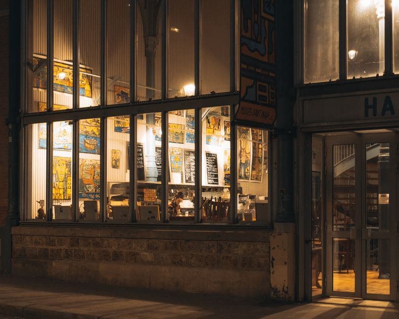 Een sfeervol verlichte horecagelegenheid met grote raampartijen en kunst aan de muur, passend bij een coffeeshop.