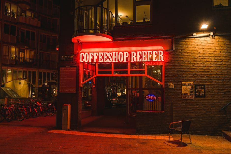 Verlichte entree van een Nederlandse coffeeshop bij nacht met een rood neonbord en geparkeerde fietsen.